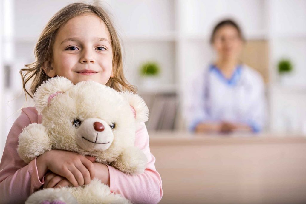 Niña abrazando un peluche con una doctora al fondo, simbolizando el cuidado infantil durante el tratamiento con antibióticos. Antibioticos en niños.