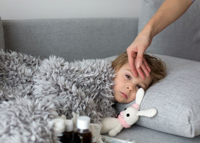 Niño enfermo en la cama con un peluche de conejo y su madre tocándole la frente; se ven frascos de medicamentos antibióticos en la mesita. Antibioticos en niños.
